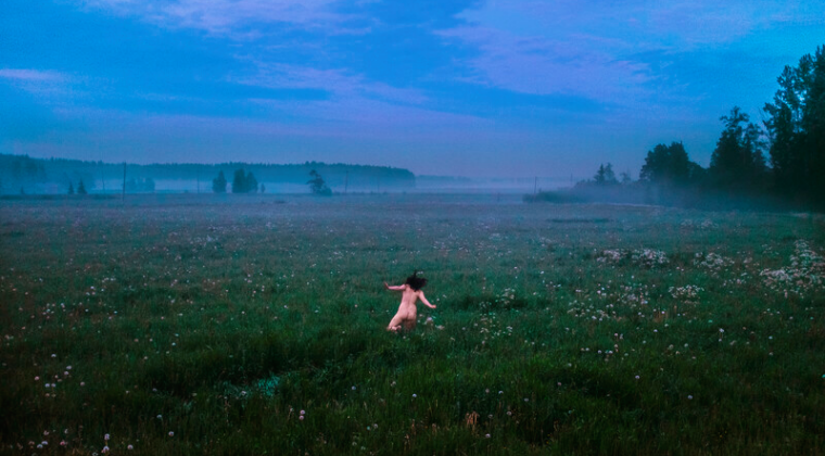 Valoisassa kesäyössä alasto nainen juoksee pellolla kukkaseppele päässä.